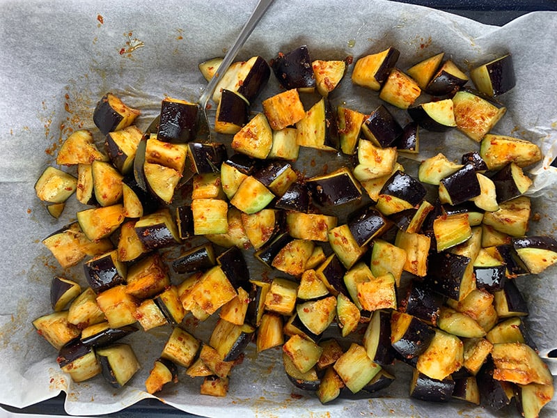 Tossing cubed eggplant with oil and harissa on a lined baking tray. 