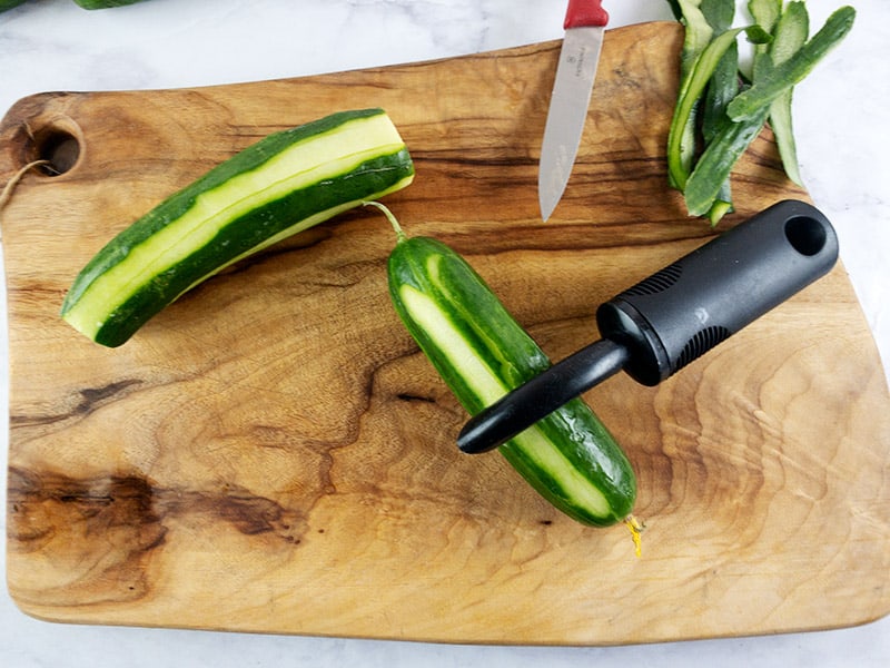 Cucumbers being peeled with a peeler on a wooden board. 
