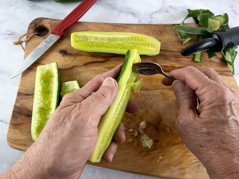 Hands deseeding a cucumber with a spoon over a wooden board.