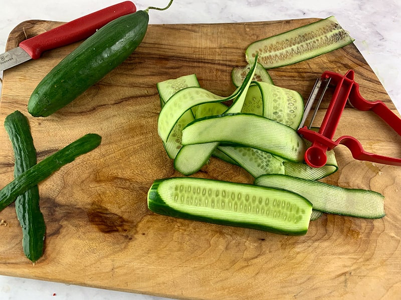 Cucumbers being sliced into ribbons with a peeler on a wooden board. 