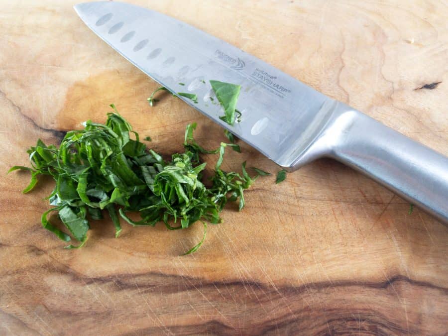 Basil leaves on a wooden board cut into thin ribbons, chiffonade style with a knife on the side. 