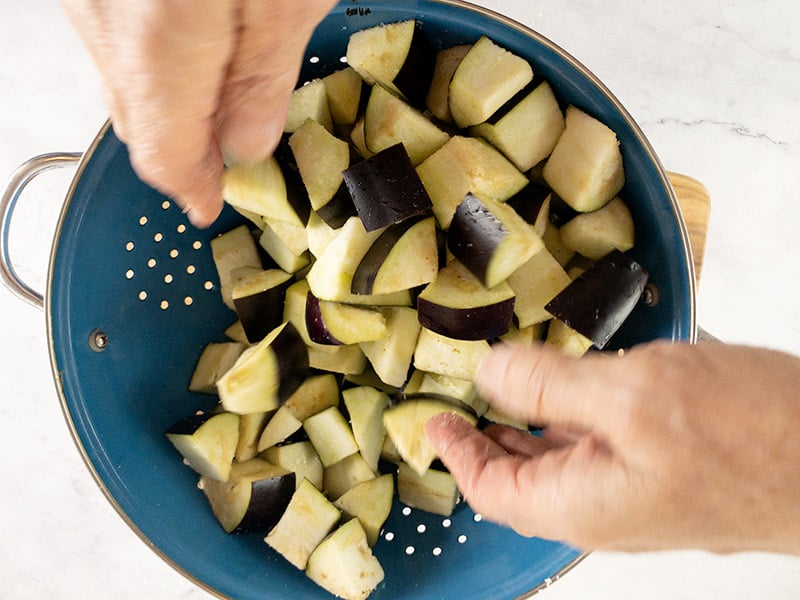 Hands Tossing eggplant cubes with salt in a colander.