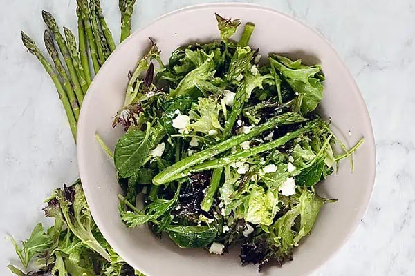 Blanched asparagus with feta salad in a pale pink bowl with salad greens and asparagus on the side.