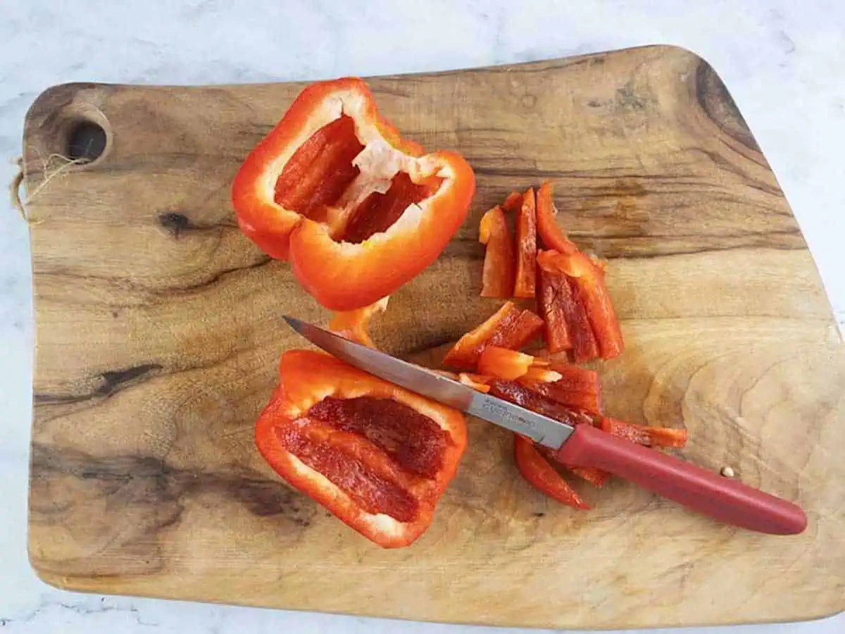 Cutting red pepper into small strips on a wooden board with a serrated knife.