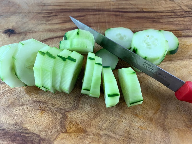 A knife cutting peeled cucumbers into rounds on a wooden chopping board.