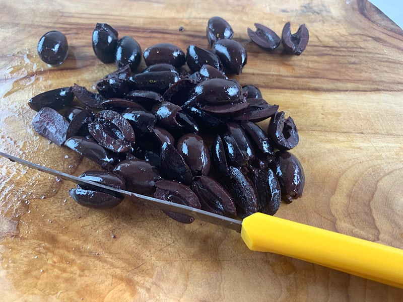 A yellow knife slicing black olives on a wooden board. 