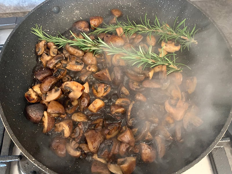 Adding rosemary and garlic to a pan with hot sauteed mushrooms.