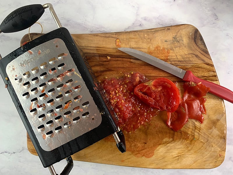Grating a ripe tomato with a box grater on a wooden board. 