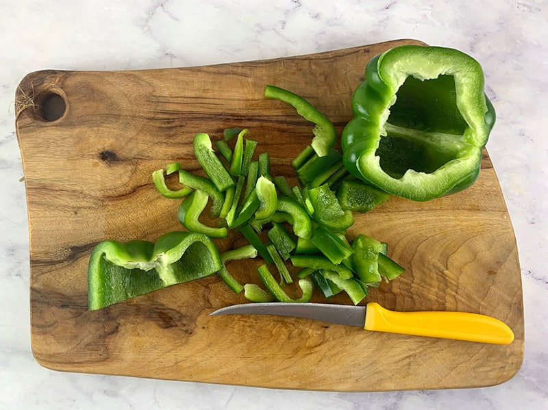 A yellow knife slicing a green capsicum or bell pepper into strips on a wooden board. 
