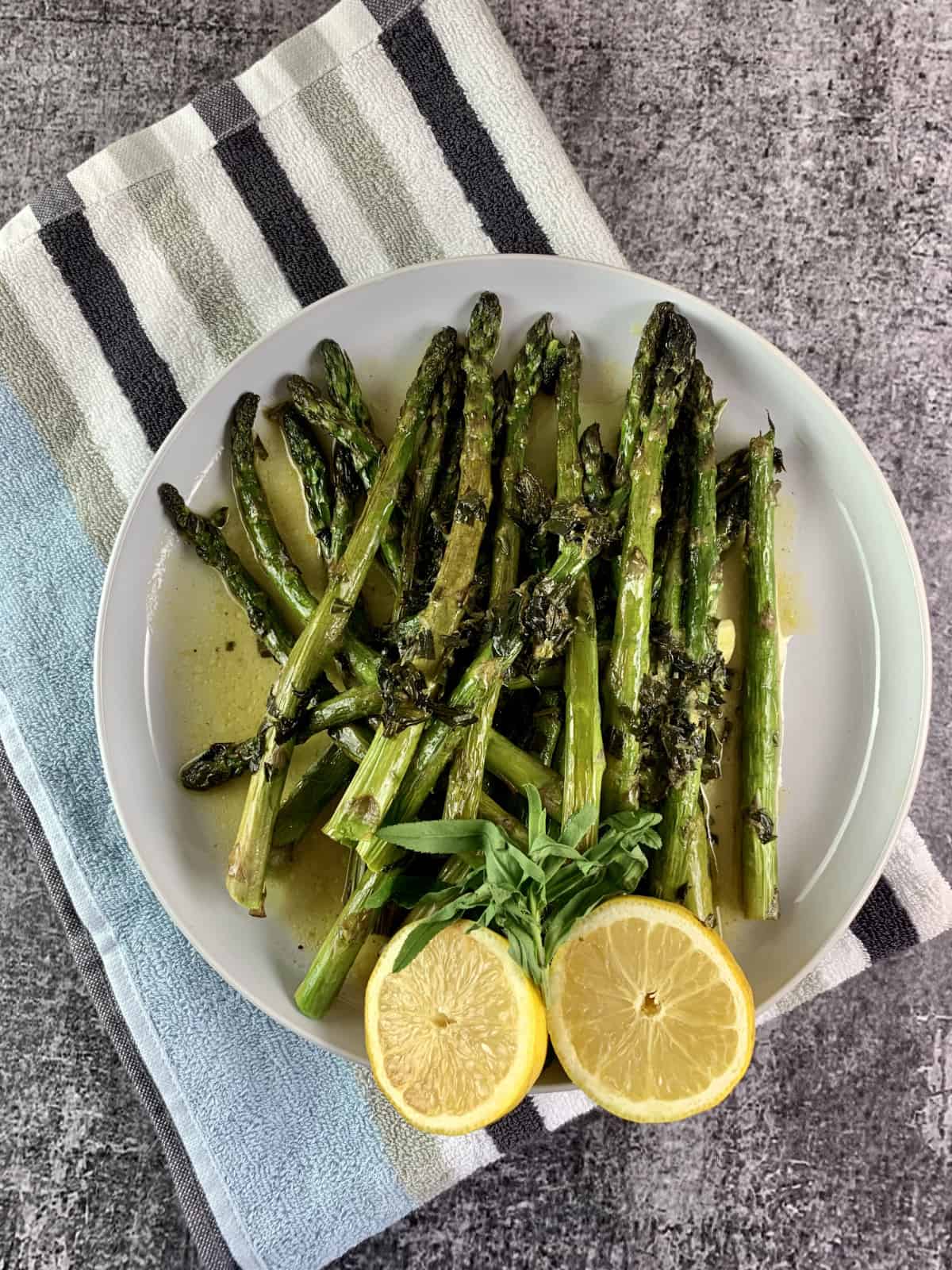 Oven roasted asparagus, with a halved lemon on a white plate on top of a striped tea towel.
