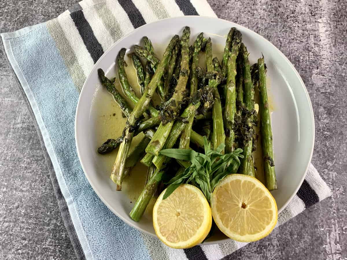 Oven roasted asparagus, with a halved lemon on a white plate on top of a striped tea towel.