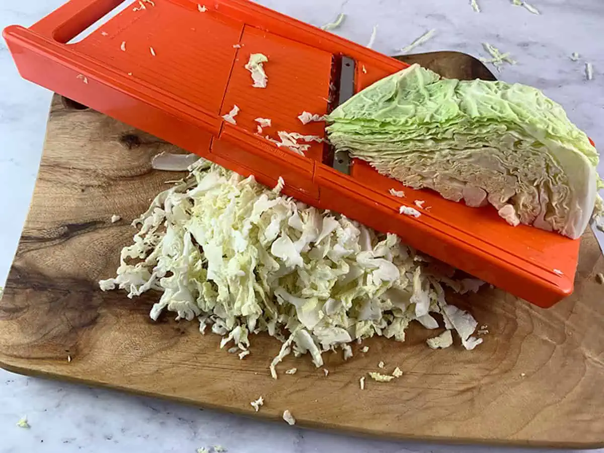 A savoy cabbage being shredded with a mandoline slicer on a wooden board.