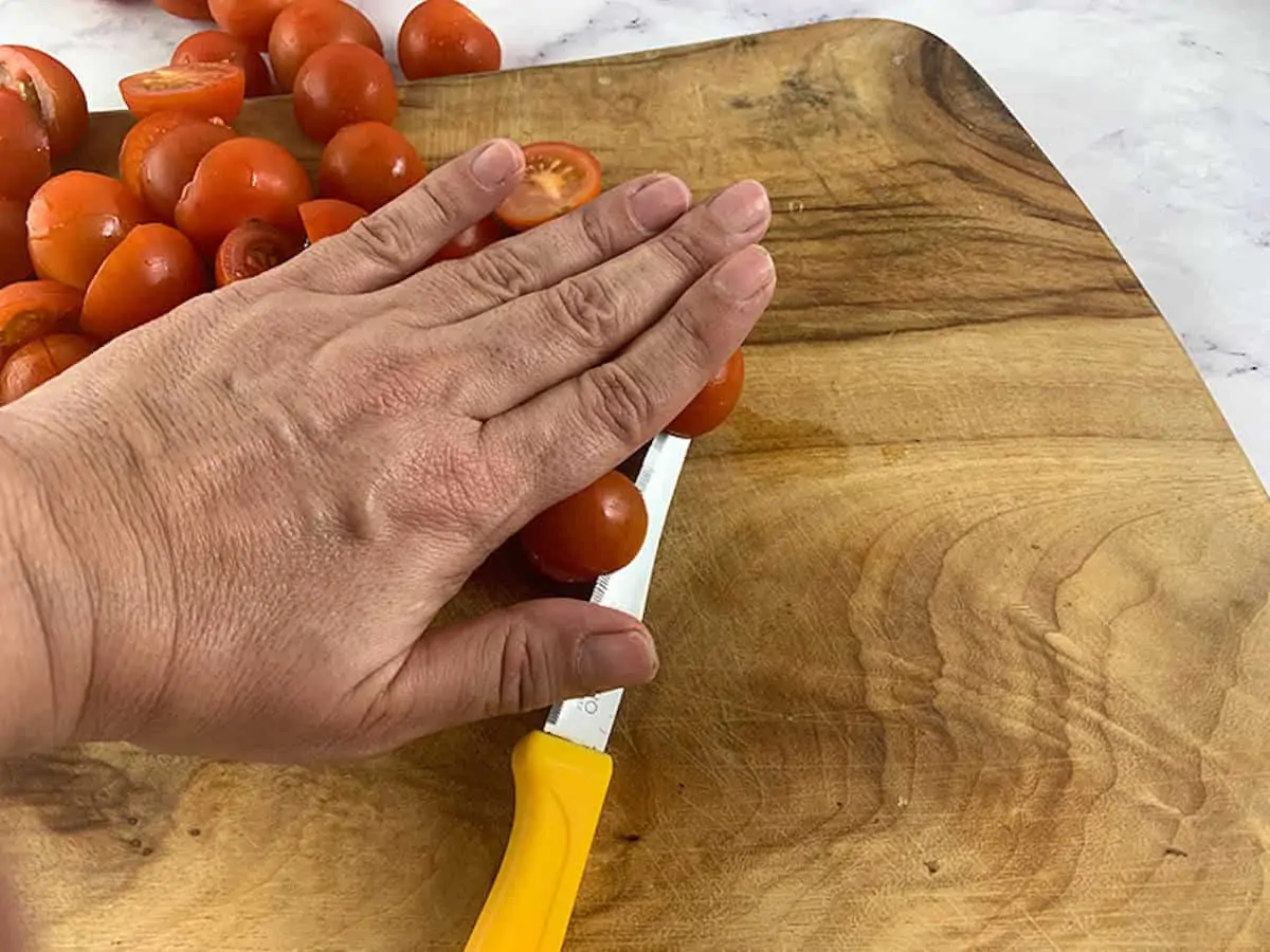 A hand on top of a pile of cherry tomatoes holding them together so that they can be sliced with a yellow knife on a wooden board.