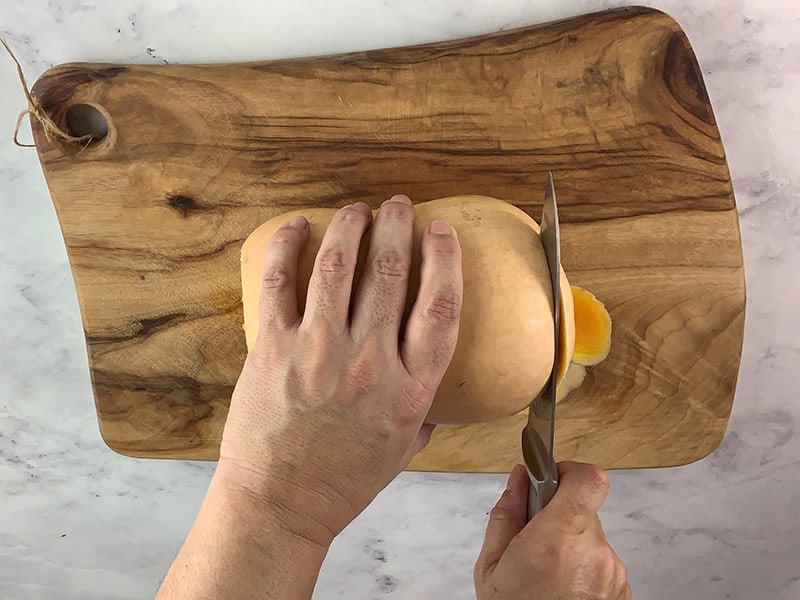 Hands trimming a butternut pumpkin on a wooden board with a knife. 