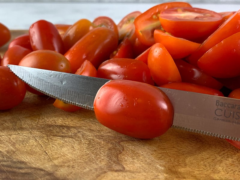 A close-up of mini Roma tomatoes being sliced in half with a knife on a wooden board. 