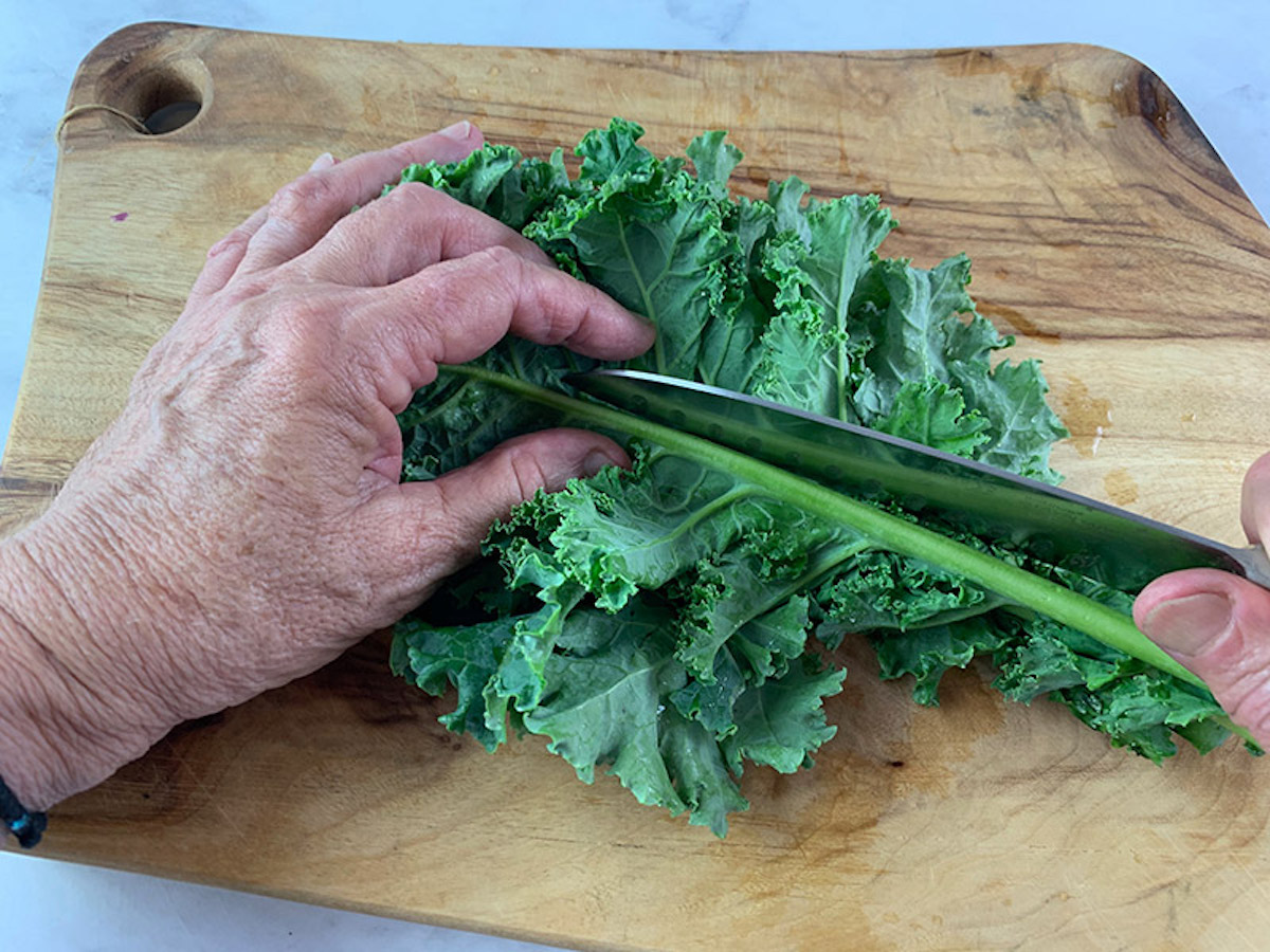 Hands cutting kale stems on a wooden board with a knife.