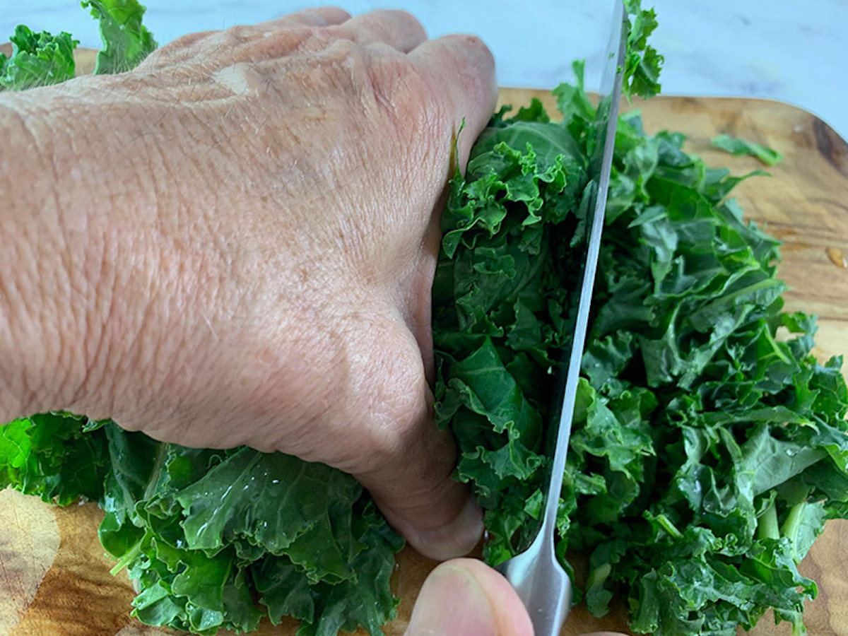 Hands shredding kale leaves with a knife on a wooden board.