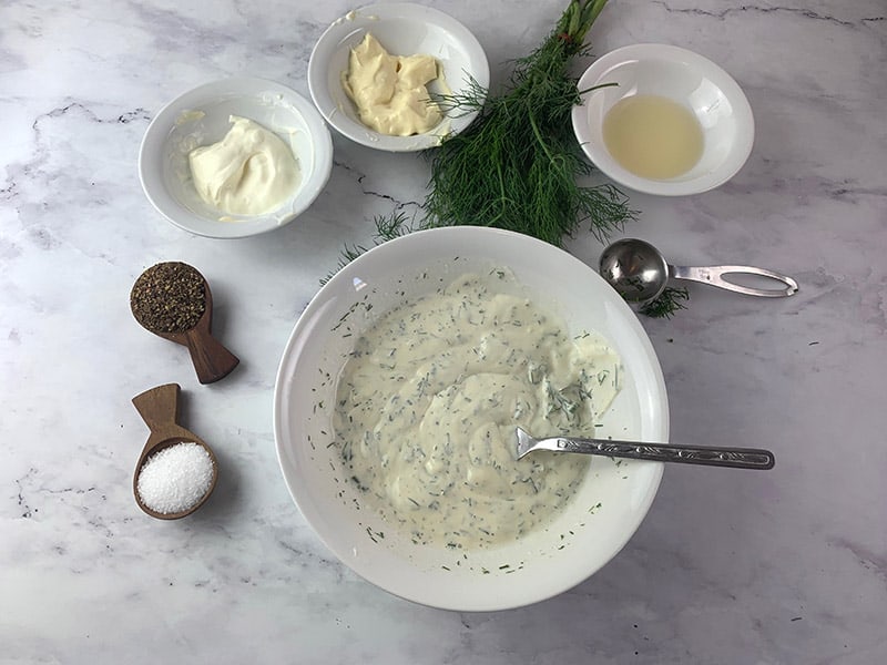 Mixing dill and yoghurt dressing in a white bowl with a spoon with ingredients scattered around. 
