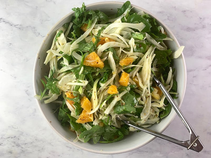 Fennel and orange salad in a white bowl that is being mixed with some tongs. 