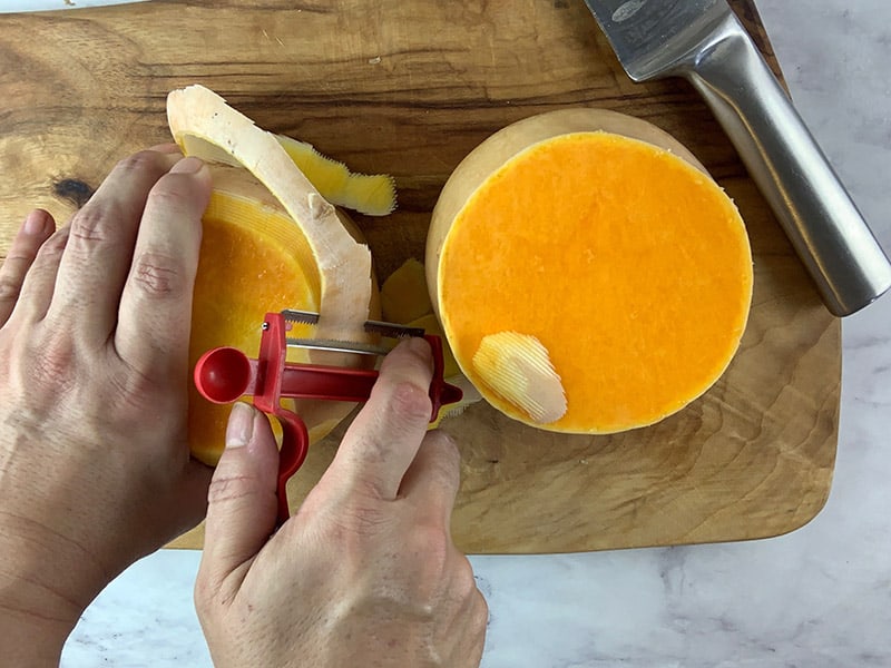 Hands peeling two butternut pumpkin halves with a peeler on a wooden board. 