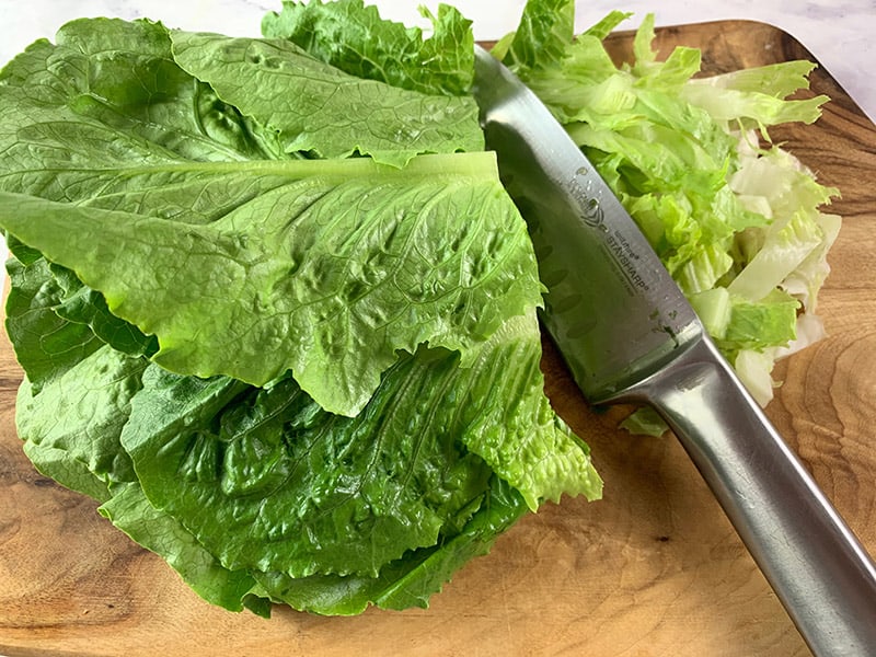 A close-up of a knife shredding cos or romaine lettuce on a wooden board. 