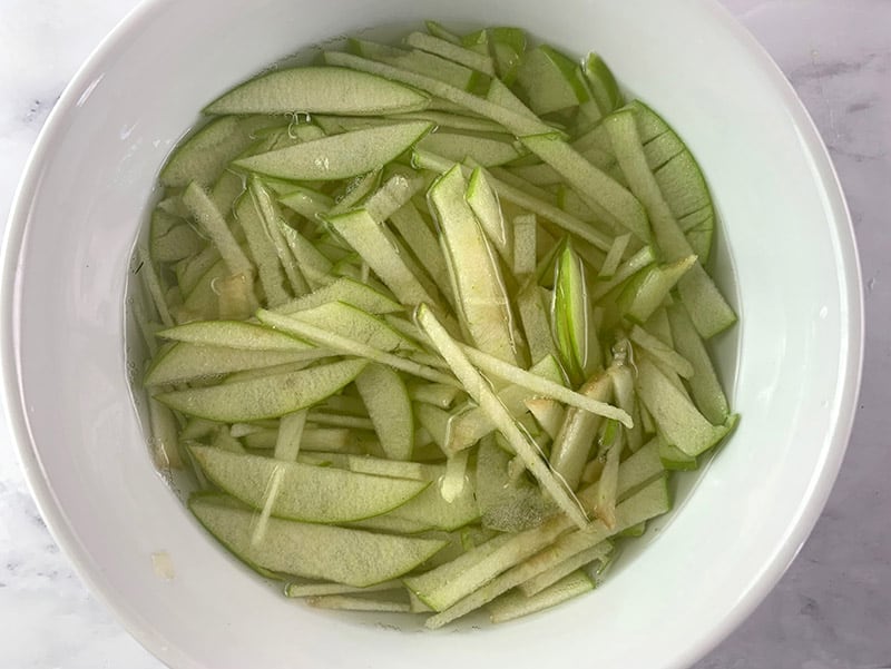 Julienned apple slices sitting in water in a white bowl to prevent growing. 