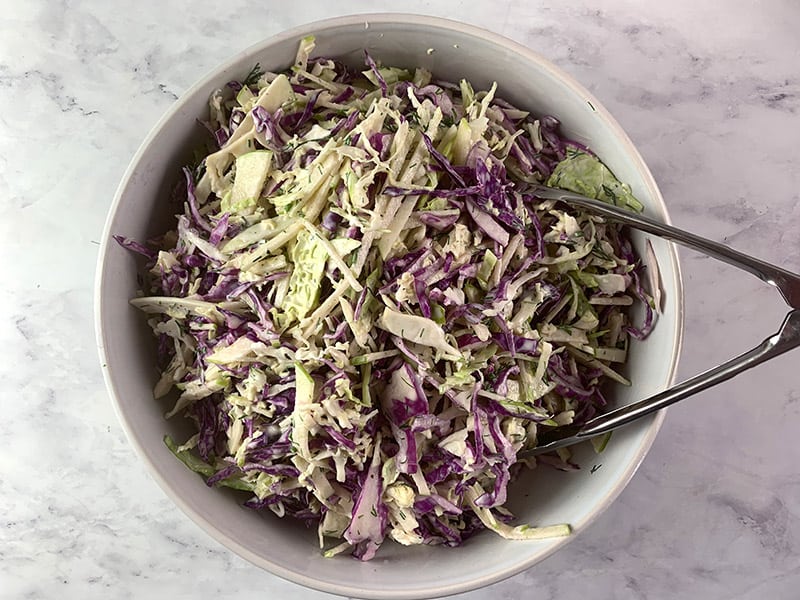 Mixing apple slaw ingredients with yoghurt dill dressing in a white bowl with tongs.