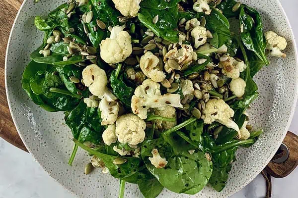 Roast cauliflower salad on a ceramic plate sitting on a wooden board with mixed seeds in a small bowl on the side.