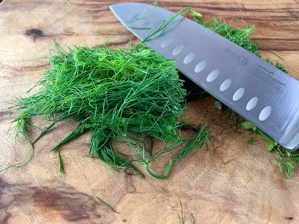 Chopping fennel fronds on a wooden board with a knife.