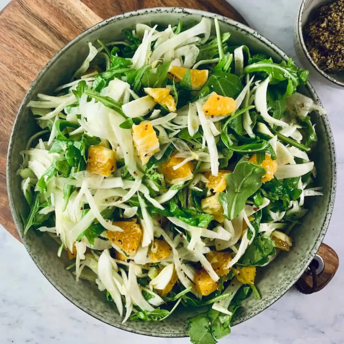 Fennel and Orange Salad in a deep ceramic bowl on a wooden board with mustard in a bowl on top right.