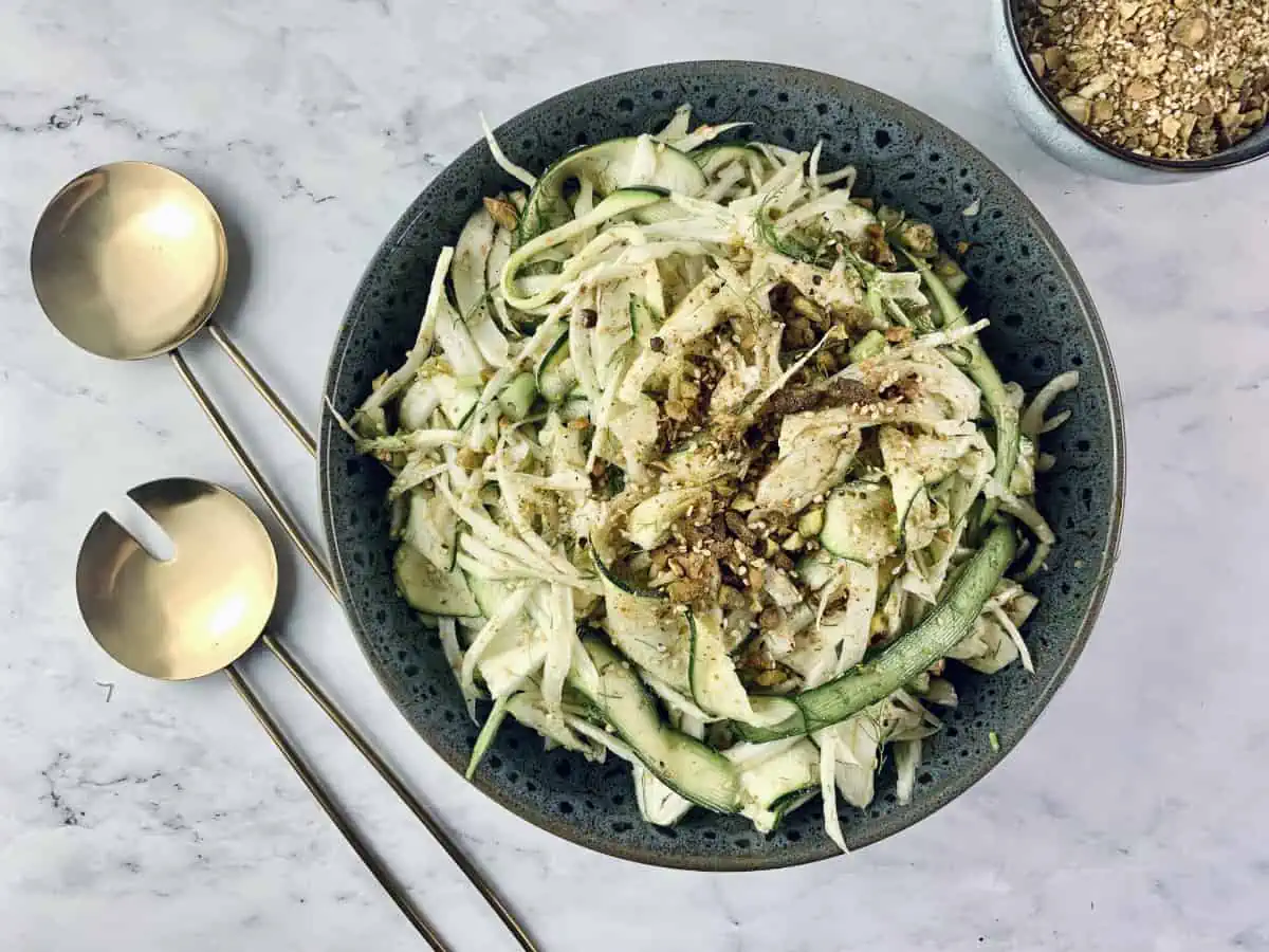 Fennel zucchini salad in a dark grey plate with gold servers and Dukkah on the side.