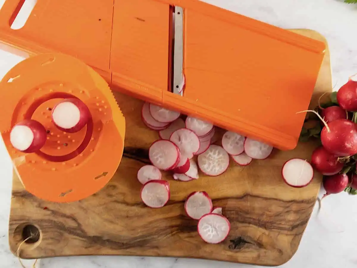 Slicing radishes with an orange vegetable slicer on a wooden board with a bunch of radishes on the side.