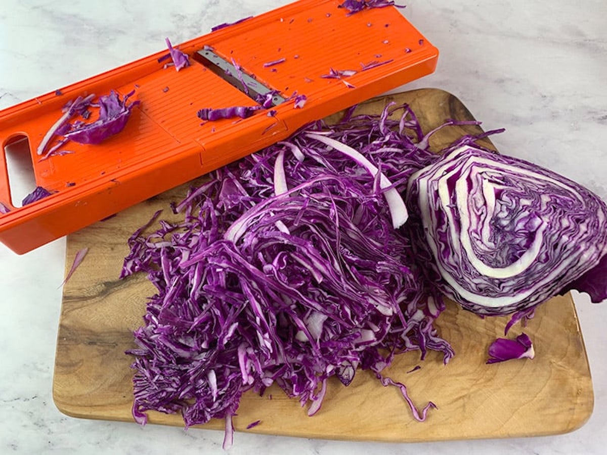 Red cabbage being shredded with a mandoline slicer on a wooden board.