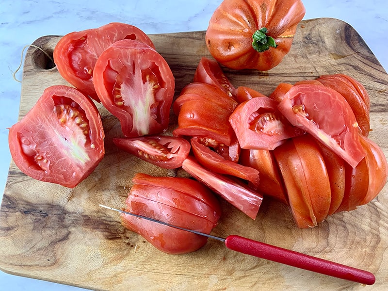 CUTTING HEIRLOOM TOMATOES INTO WEDGES ON A WOODEN BOARD WITH A SMALL RED SERRATED EDGE KNIFE