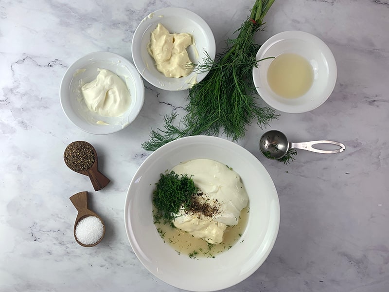 Ingredients for creamy dill dressing in a bowl with a bunch of dill, lemon juice, yoghurt, mayo, salt and pepper on the side.