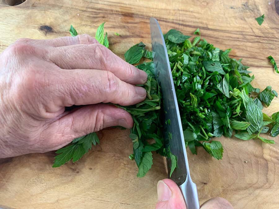 Hands roughly chopping herbs on a wooden board with a knife.