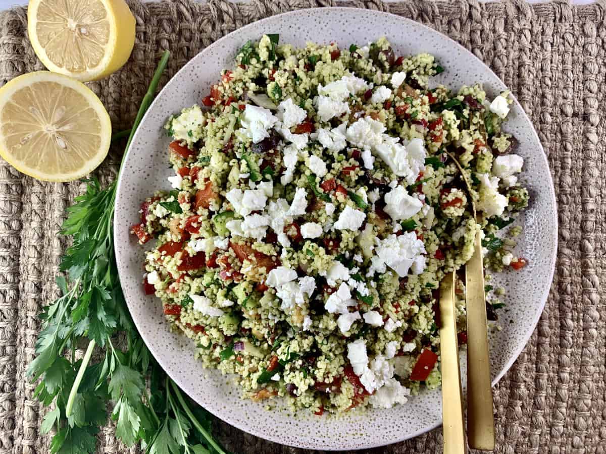Mediterranean couscous salad on a ceramic plate with gold servers and sliced lemons and parsley on the side.