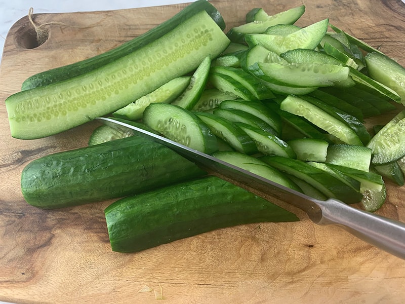 A knife slicing cucumbers on the diagonal on a wooden board.