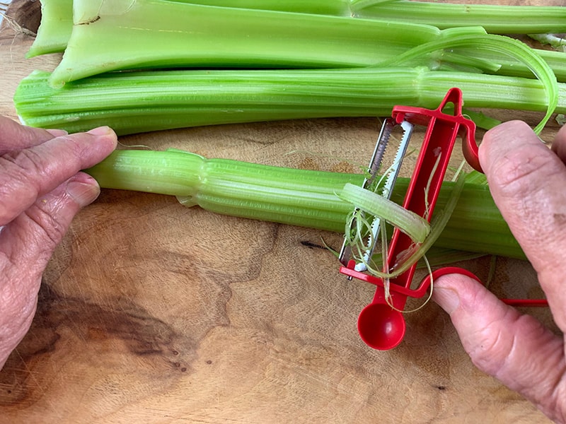 DE-THREADING CELERY STALKS WITH A VEGETABLE PEELER ON A WOODEN BOARD