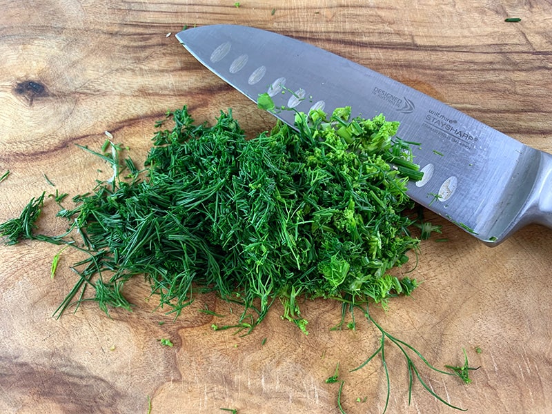THINLY CHOPPING DILL WITH A STEEL KNIFE ON A WOODEN BOARD