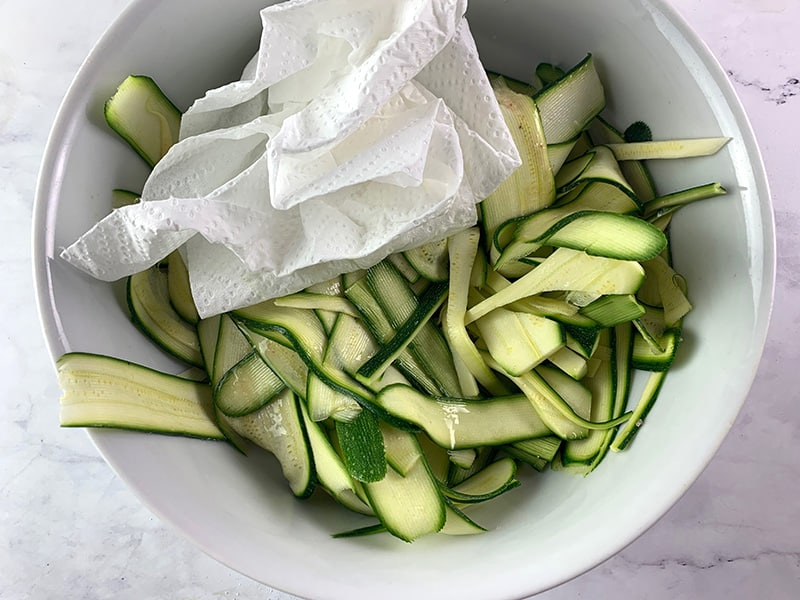 Salted zucchini ribbons on a plate being patted down with a paper towel.