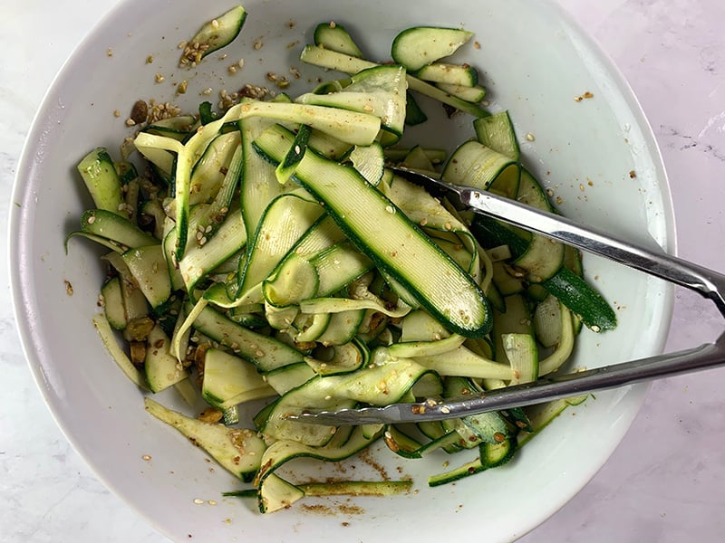 Mixing zucchini ribbons with seasonings in a white bowl with tongs.