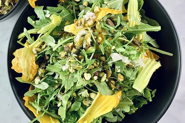 Arugula salad with squash flowers in a black bowl with pistachios in a small black bowl and zucchini flowers on the side.