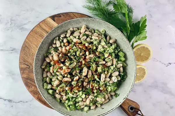 White bean salad in a ceramic bowl sitting on a wooden board with celery stalks and lemon halves on the side.
