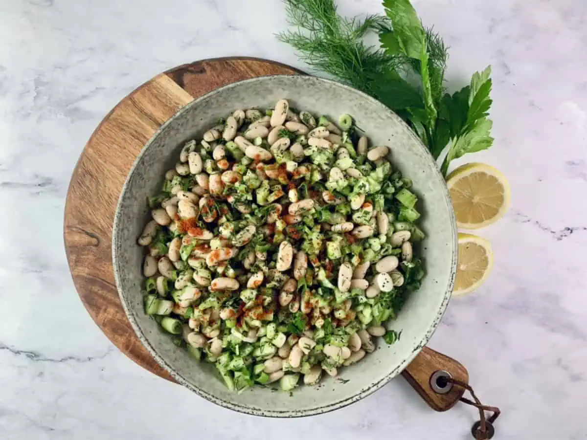 White bean salad in a ceramic bowl sitting on a wooden board with celery stalks and lemon halves on the side.