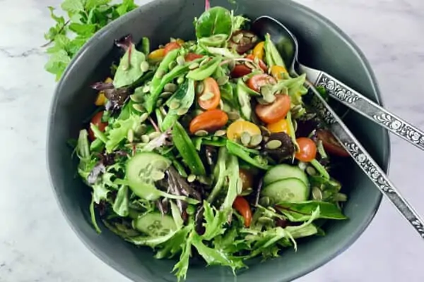 Spring Mix Salad in a dark grey bowl with mint springs on the top left corner and silver salad servers on the right.