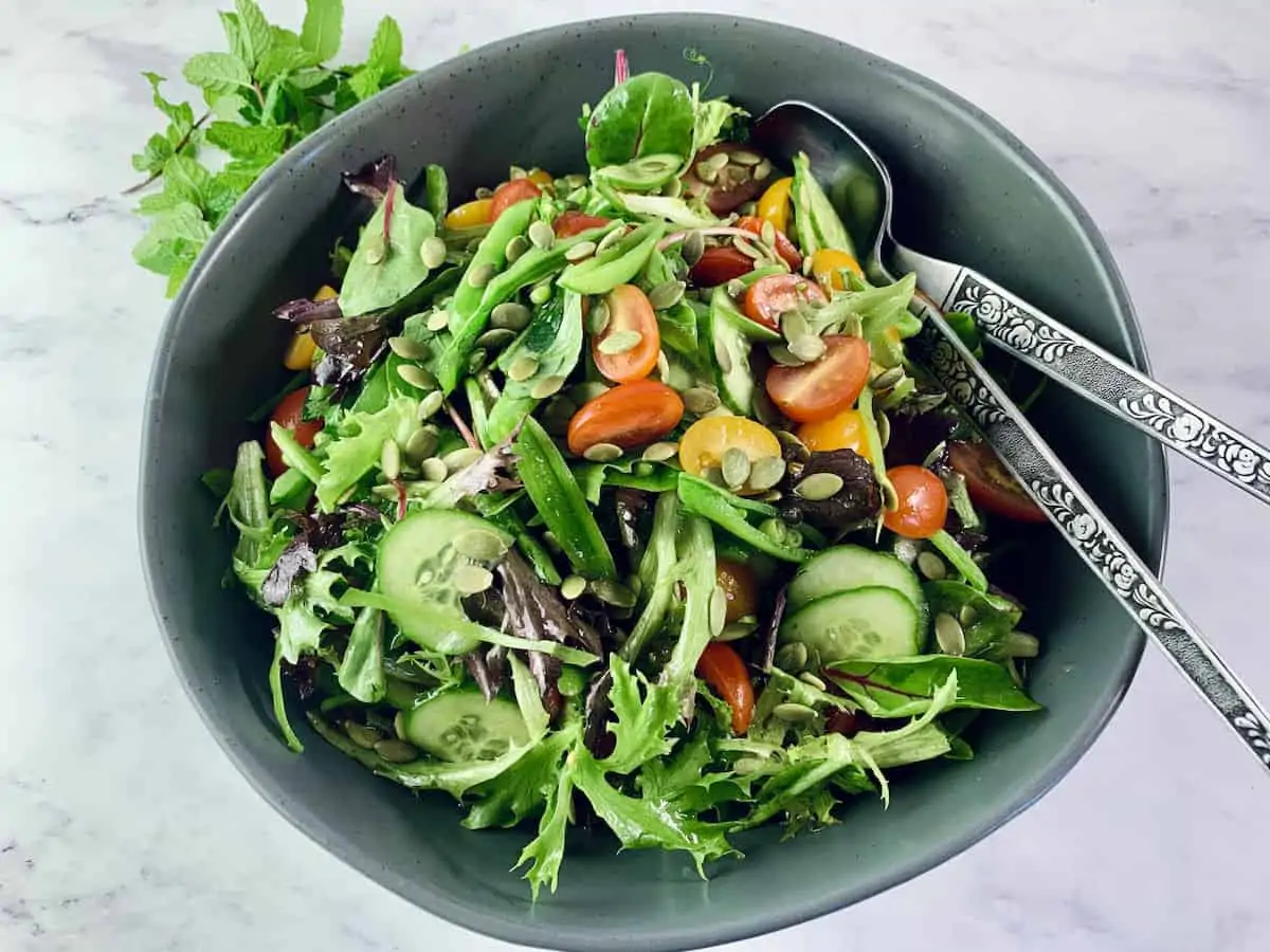 Spring Mix Salad in a dark grey bowl with mint springs on the top left corner and silver salad servers on the right.