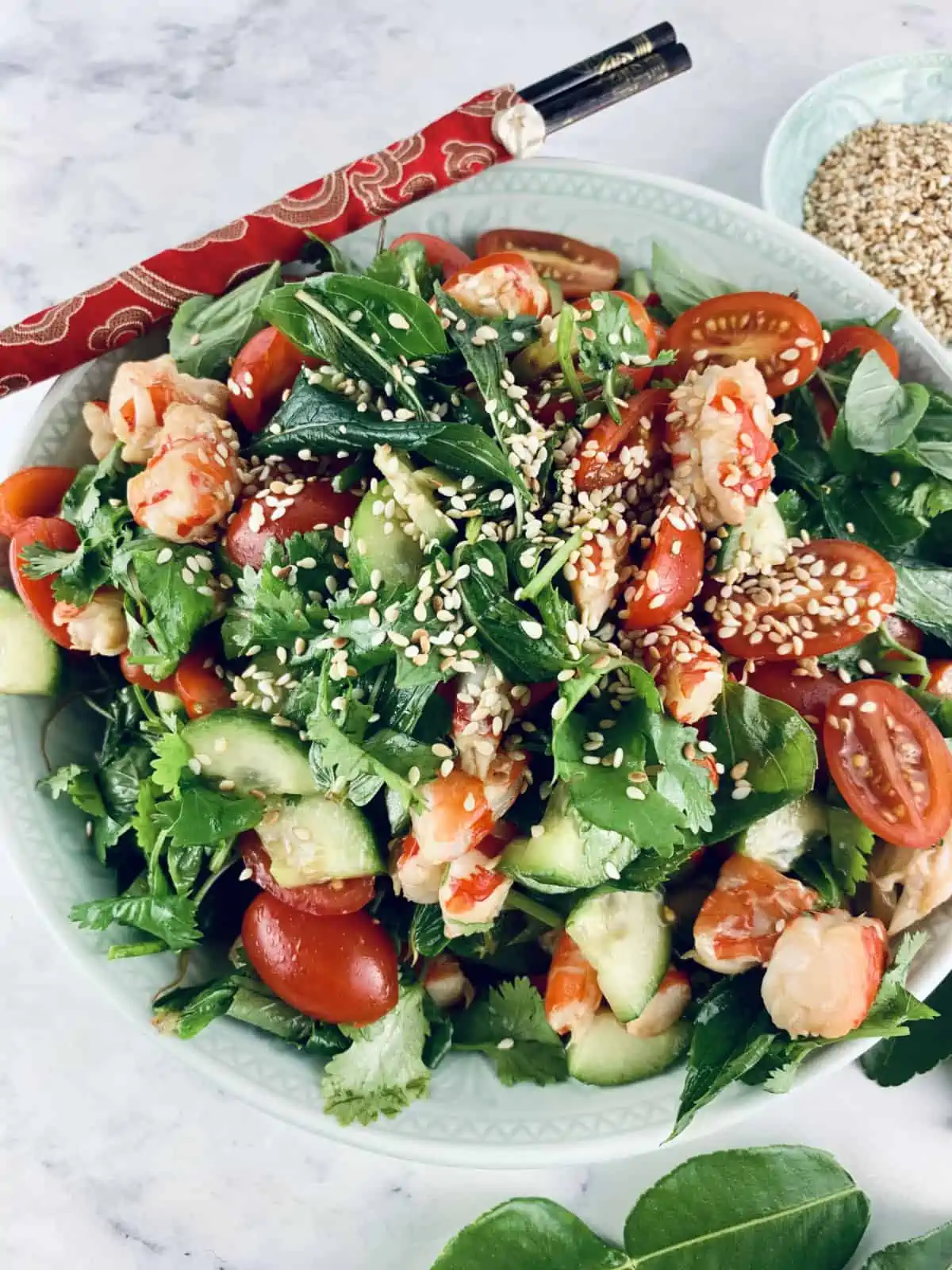 Thai Prawn Salad in a mint bowl with red chopsticks in the top left corner, sesame seeds in the right corner and lime leaves at the bottom.