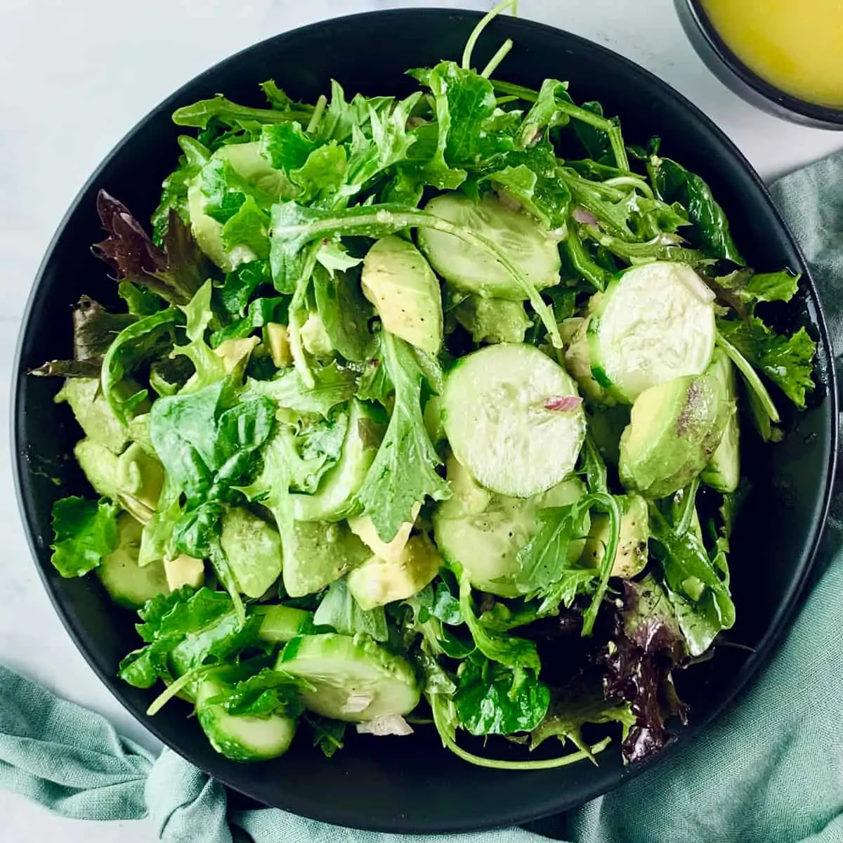 French green salad in a black bowl with a mint linen napkin at the bottom and dressing in a small black bowl in the top right corner.