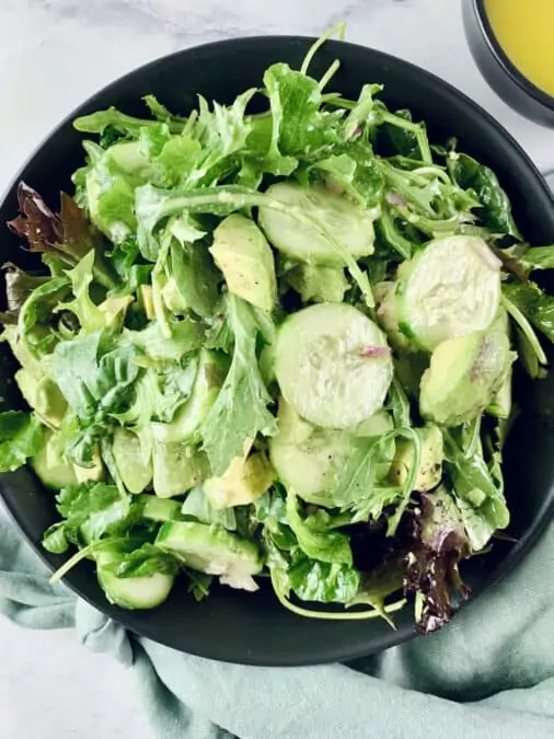 French green salad in a black bowl with a mint linen napkin at the bottom and dressing in a small black bowl in the top right corner.